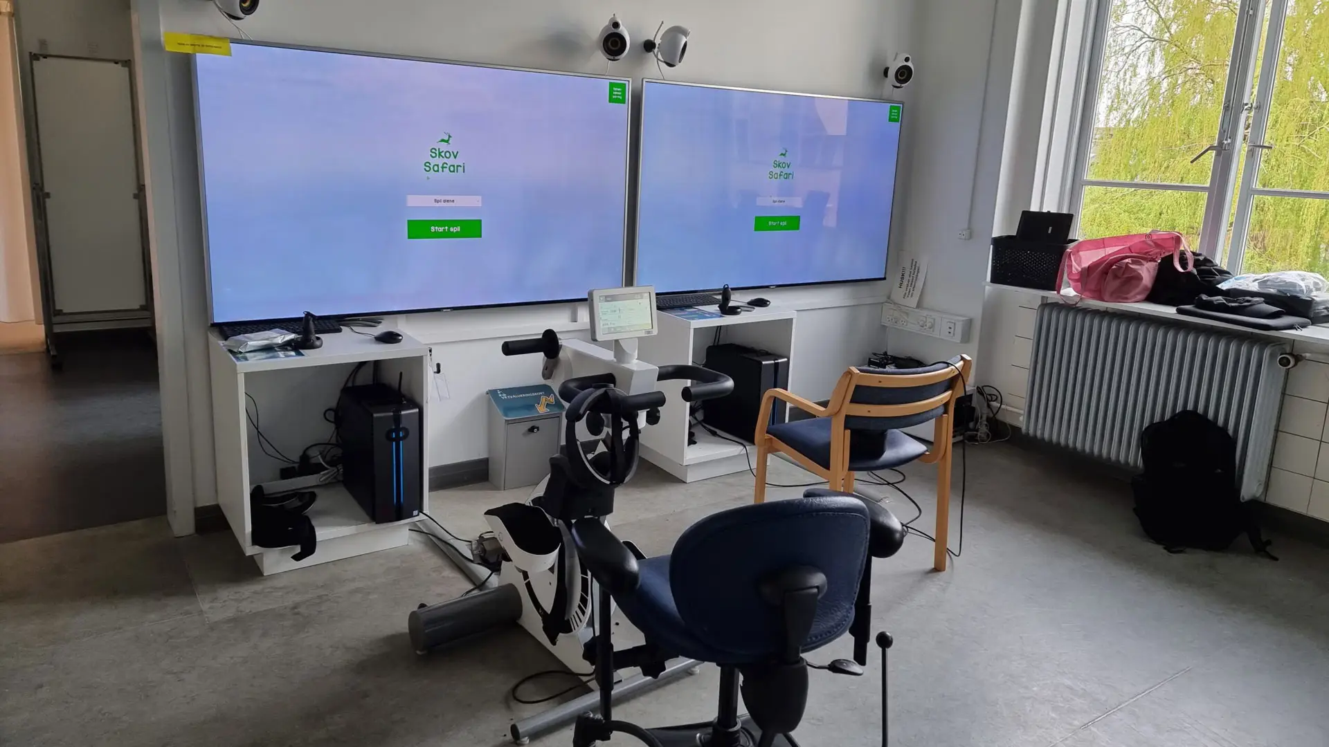 Experimental setup at Frederiksberg Hospital showcasing the VR Multiplayer Wildlife Biking project with stationary bikes and dual large screens for gameplay.